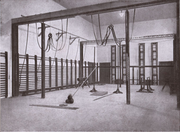 Schwarzes und weißes Foto eines Gymnastikraums mit Übungsgeräten wie Stangen und Seilen auf dem Boden, Fenstern im Hintergrund und Text unten.
