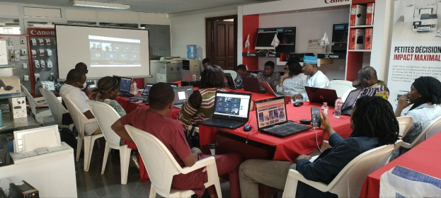 Gruppe von Menschen, die um einen Tisch mit Laptops, Flaschen und anderen Gegenständen während eines digitalen Marketing-Workshops sitzen, mit einem Banner, einem Bildschirm, einer Tür und Deckenleuchten im Raum.