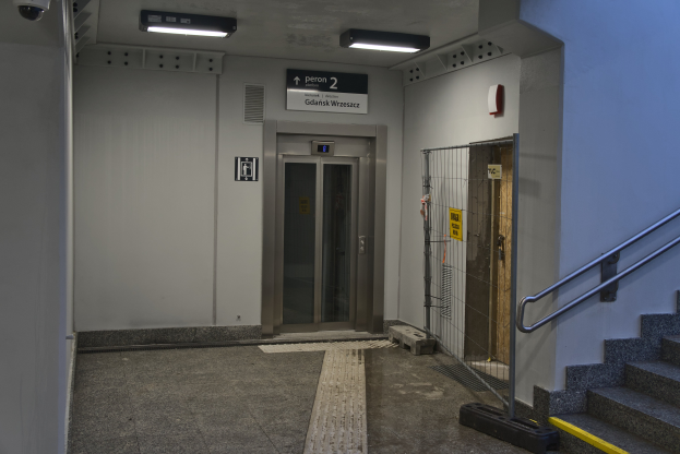 Ein Aufzug ist in einem Gebäude mit Treppe auf der rechten Seite positioniert, das eine Geländer, Beleuchtung und eine Überwachungskamera an der Spitze hat; im Hintergrund gibt es eine Wand mit einer Tür und einer Texttafel.