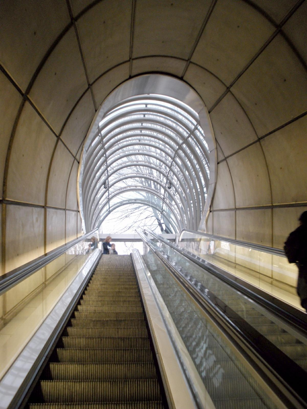 Rolltreppe in einer U-Bahn-Station mit Menschen, Geländern, Informationsschildern, Wandlampen und einem Einkaufswagen unten.