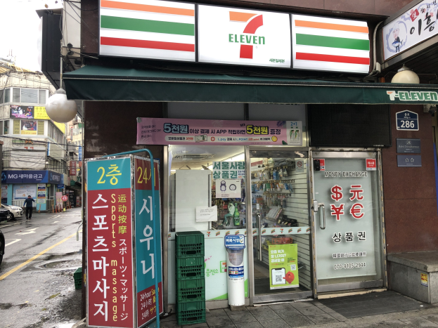 Ein 7-Eleven-Konvenienzladen in Seoul, Südkorea, mit Fahrzeugen auf der Straße, einem Fußgänger auf der linken Seite und Gebäuden im Hintergrund unter einem sichtbaren Himmel.